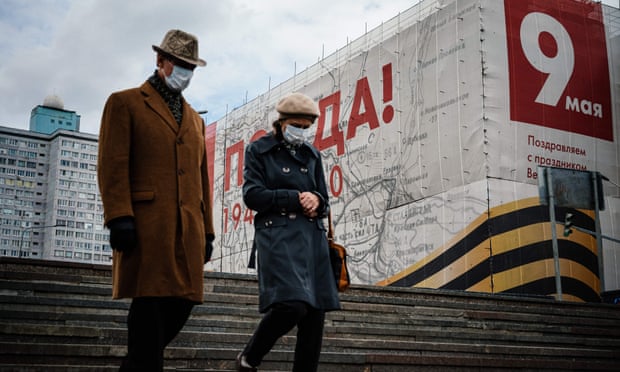 A couple wearing face masks walk in front of a banner for the upcoming Victory Day celebrations
