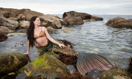 Hannah Cadec, comme la sirène Taurva, avec une queue brune et dorée, assise sur un rocher dans la mer
