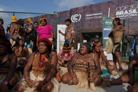 A group of Indigenous women with tribal tattoos and grass skirts sit in front of a building labeled Cop30 Brasil.