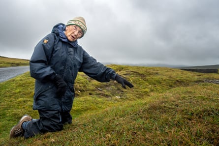 An elderly woman, Margaret Bradshaw, kneels on a grassy hillside, pointing at small flowers