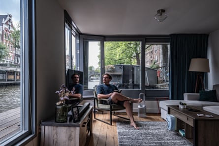 Hagen Harvey in a neat, minimalist room is sitting in a chair with a book open as he looks out the window at the water