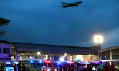 Ambulances lined up at dusk next to airport building with plane in sky above.