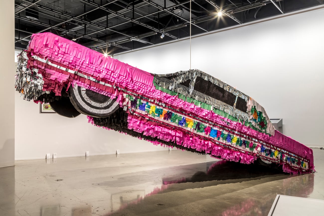 A lifesize version of the legendary pink Chevrolet Impala is recreated in paper fringe and cardboard and hung at an angle from the ceiling of an art gallery.