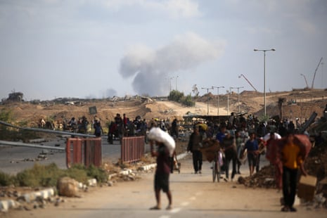 A plume of smoke rises in the background as Palestinian people return from a food distribution point in the central Gaza Strip.