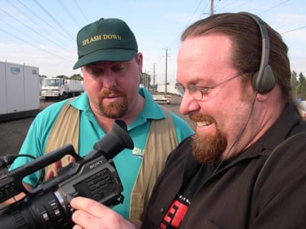 Head and shoulders photo of two men standing close together, with one of them looking into a video camera viewer and smiling, while the other looks on.