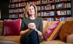 Arianna Huffington sitting forward and cross-legged on her sofa at home, cupping a mug in her hands, bookshelves lining the wall behind her