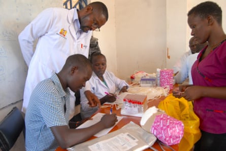 A young man holding a pen reads a document while a doctor leans over his shoulder. The table he is siting at is holds empty blood collection vials and bloodbags.