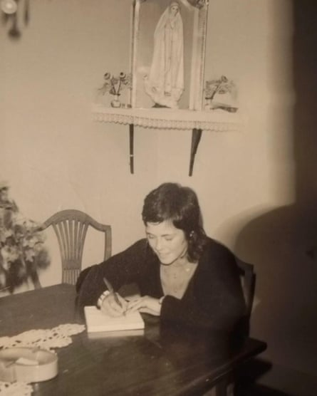 A black and white photo of a woman sitting at a table with a pen and paper and a small statue of Mary on the wall behind her