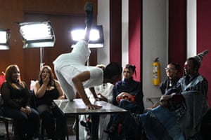 A person in a white outfit sticks their leg in the air as they crouch on top of a small table while people in seats watch and very bright studio lights glow in the background