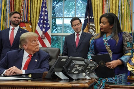 Thérèse Kayikwamba Wagner (right) stands beside Donald Trump, who is seated at his desk in the Oval Office. JD Vance and Marco Rubio are stood behind