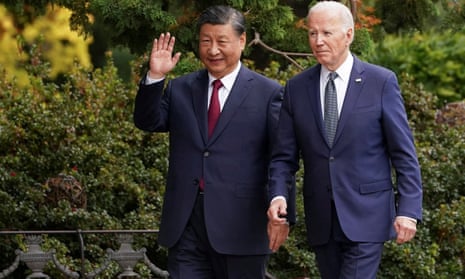 Two men in navy suits and ties walk in front of green bushes