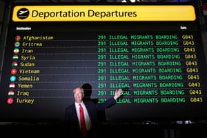 A man in a suit points to a large mock departures board behind him showing ‘illegal migrants boarding’ flights to countries including Afghanistan and Eritrea