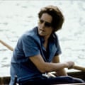 Hugh Grant’s character rowing in Stoke Park’s lake. Grant wears aviator sunglasses, a blue shirt, and a cigarette hands from his lips. 