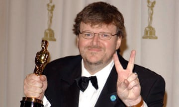 Man wearing glasses, black suit and black bowtie holds a gold statue and gestures peace sign