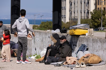 People with bags and suitcases sitting or lying against a wall with the sea in the background.