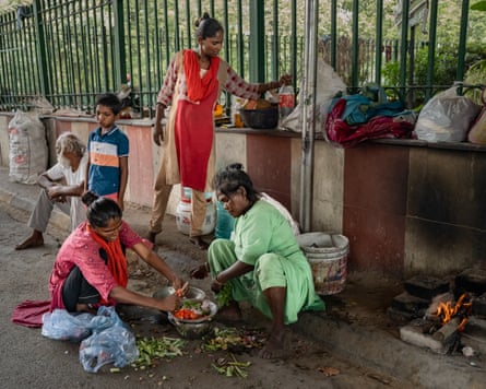 Two women crouch over metal bowls preparing vegetables while another woman stands to place a plastic bottle of water next to the railings behind them.