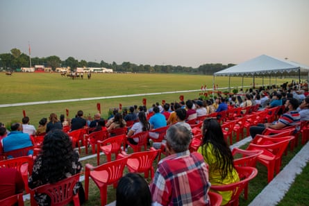 People on rows of chairs by the side of a big expanse of grass watching a polo match in the distance