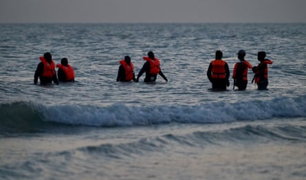 Seven people of varying heights, seen in near-silhouette walk through the waves and shallow water towards the sand. They wear bright orange lifevests.