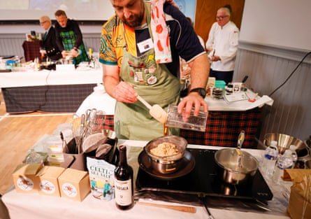 Rob Reid spoons oats into a glass bowl on a small portable induction hob on a tabletop. Ingredients are lined up in front of him including a packet of sea salt and a bottle of marsala. He wears a green apron and a Wallabies rugby shirt.