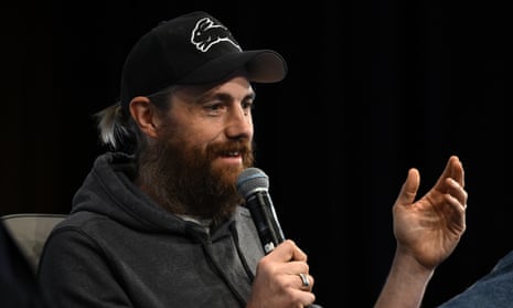 Mike Cannon-Brookes, wearing a grey hoodie and a black cap, speaks into a microphone and gestures with one hand