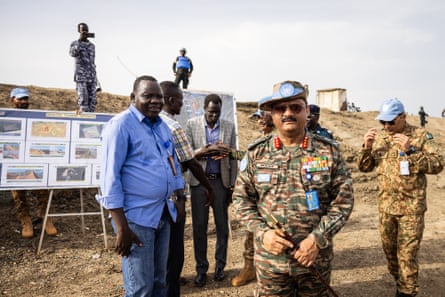 A group of men, some in suits and some in military fatigues, stand in front of a board which reads ‘Bentiu flood update’. Subramanian stands closest to, and facing, the camera.