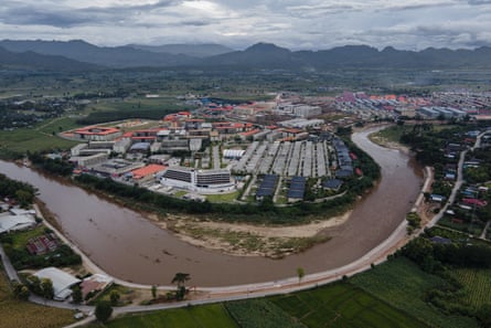 Aerial view of a sprawling compound on a lush plain by a large river with prison-like accommodation blocks, flats, offices and hangars surrounded by a perimeter fence