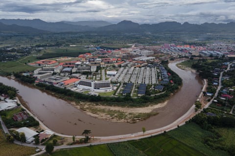Aerial view of a sprawling compound on a lush plain by a large river with prison-like accommodation blocks, flats, offices and hangars surrounded by a perimeter fence