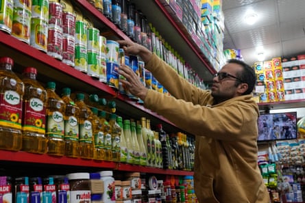 A shopkeeper arranges items on a shelf in a shop, including bottles of cooking oil