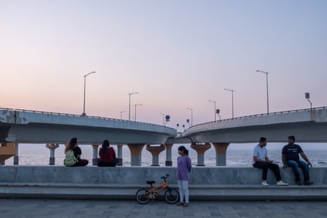 Two couples sitting on a sea wall, while a woman stands in the middle by a bicycle, with two lanes on piles curve away in the background