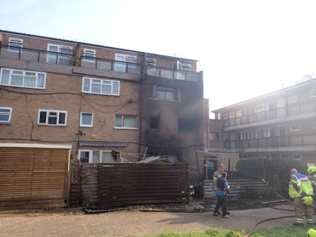 Fire damage including charred brickwork and burned-out windows at a square, modern four-storey block of flats. Firefighters stand in front of the property with hoses.