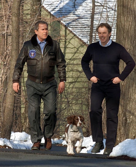 George W Bush, wearing a leather jacket, a blue shirt, grey trousers and brown shoes, and Tony Blair, wearing a blue shirt and a black jumper and trousers, walk on a path with snow beside it, accompanied by Bush’s springer spaniel Spot