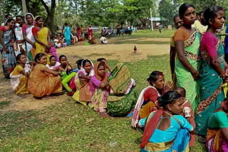 A line of women in colourful saris snakes through a park with some sitting down on the grass as they wait