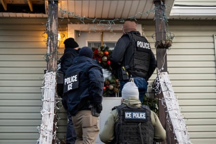 Four men wearing bullet-proof vests with ‘Police – ICE’ written on the back stand on the front steps of a house with a Christmas wreath on the door