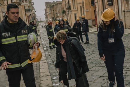 An elderly woman walks with head bowed along a cobbled street between houses, flanked by firefighters