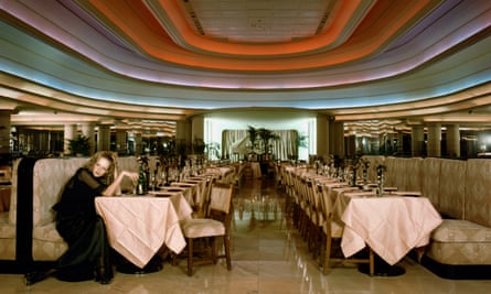 Twiggy sits alone at a table in a room with polished marble floor. Rainbow-coloured lighting is on the ceiling above