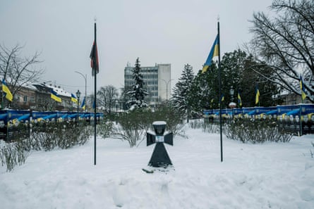 A cross that appears to be made from stone between two flags in the snow. Either side of the flags is a line of boards carrying portraits of men.