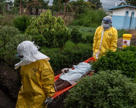 Two people wearing full personal protective equipment carry a person on a stretcher through hospital grounds.