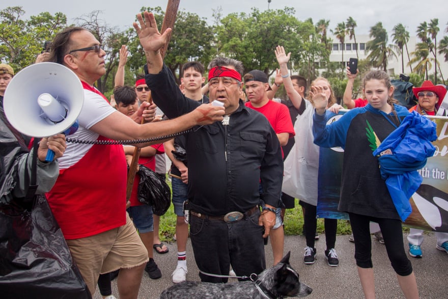 A group of people from the Lummi Nation stand together with their hands raised in protest. A man in front sings into a loudspeaker held by another man.