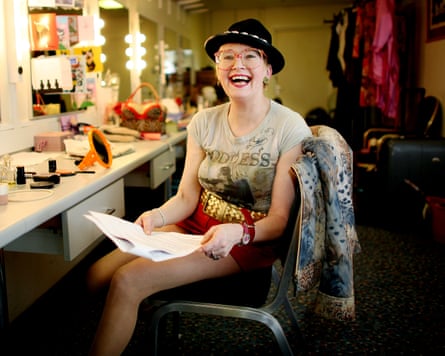 A woman with a black hat on, colourful glasses and red shorts sits laughing in a dressing room.