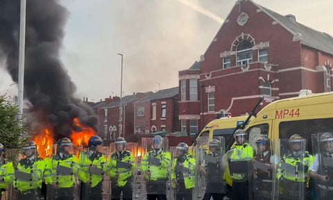 A fire burns behind a line of police officers in riot gear