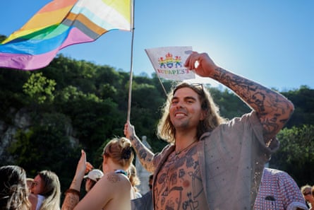A tattooed man smiles at the camera holding a LGBTQ+ flag