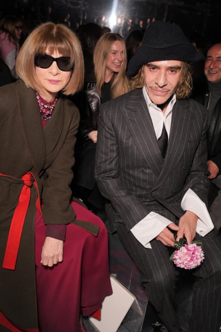 A woman in sunglasses sits next to a man wearing a suit and hat at a fashion show