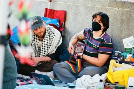 two young people sit on the ground outside, one wearing a mask and playing a guitar, the other wearing a keffiyeh