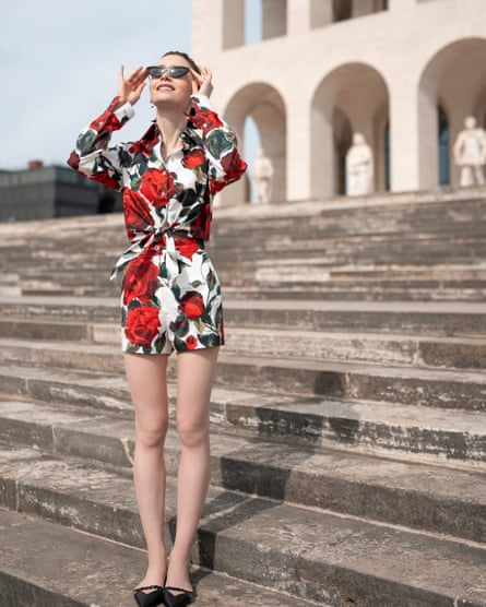 Emily stands on steps wearing a red, black and white short floral playsuit, and black cat-eye sunglasses