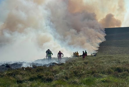 Thick smoke rising from a green heath and drifting into the sky as firefighters approach