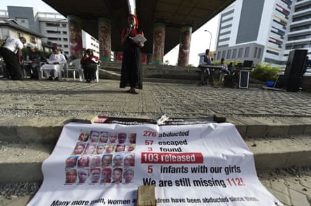 A woman speaks into a microphone under a flyover in a city, with other people sitting on chairs, behind a banner with photographs of girls