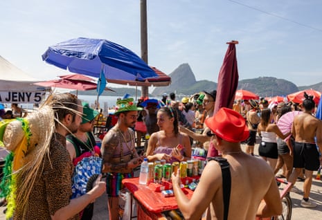 People buy drinks from a cart on a crowded beach with people standing under beach umbrellas.