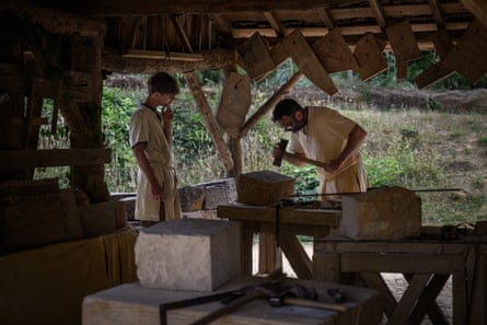 Two stone-cutters in beige, loose-fitting 13th-century garb work within the construction site of Chateau Guedelon. One watches on while the other holds a hammer above a block of stone