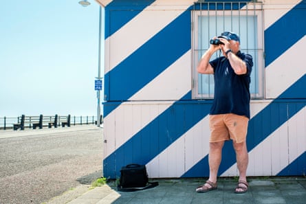 Dick Houghton wearing a navy T-shirt and salmon shorts and standing in front of a blue-and-white-striped beach building holding a pair of binoculars to the summer sky