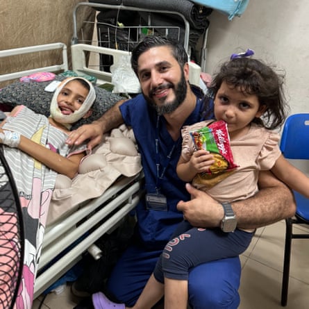 A man in surgical scrubs squats beside a bandaged girl in a bed with another child on his knee